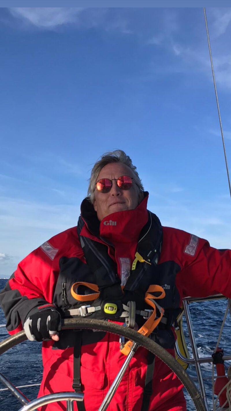 Jon Morby at the helm of a yacht, in red sailing gear and mirrored sunglasses